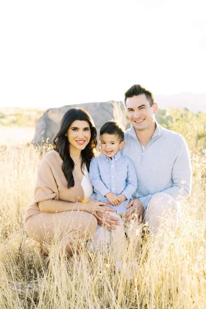 A family of three, including a woman, a man, and a young boy, pose together outdoors in a field of tall, dry grass with sunlight in the background.