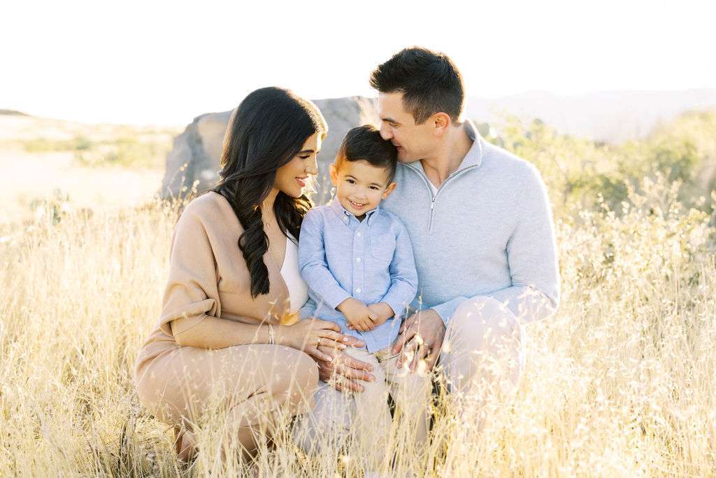 A woman, a man, and a young boy sit together in a sunny field of tall grass, smiling and looking at each other.