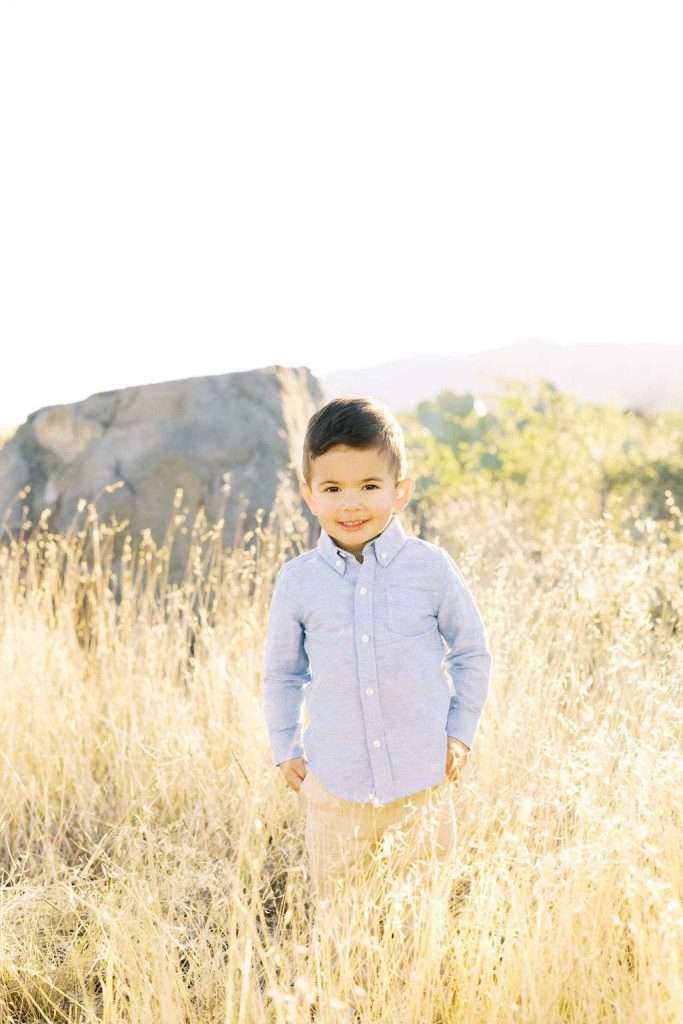 A young boy in a light blue shirt stands in a sunlit field of tall, dry grass with a large rock and trees in the background.