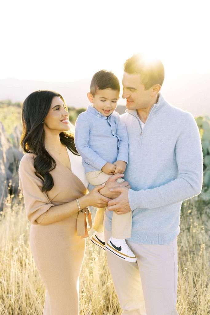 A woman, man, and young boy stand together outdoors in a field, smiling at each other with sunlight in the background.