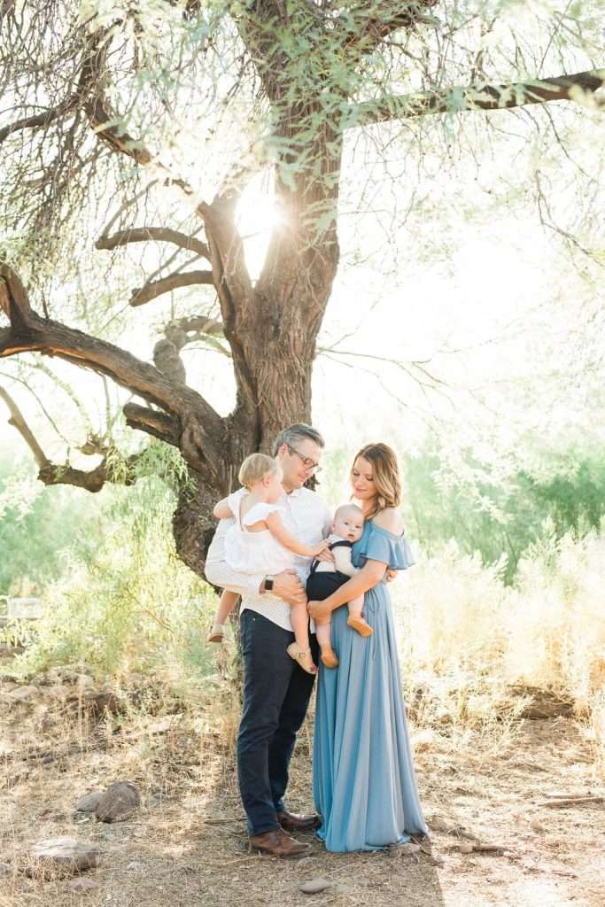 A man and woman stand outdoors with two young children near a large tree, surrounded by sunlight and greenery.