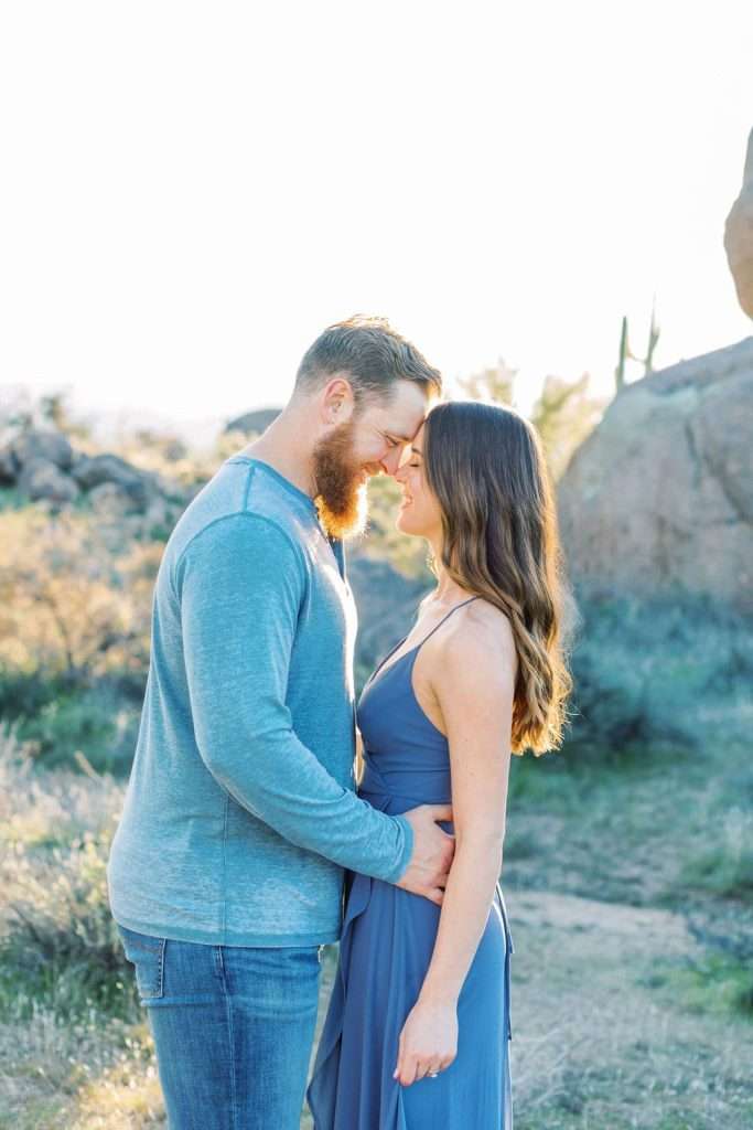 A man and woman stand close together outdoors, touching foreheads and smiling, with sunlight and desert vegetation in the background.