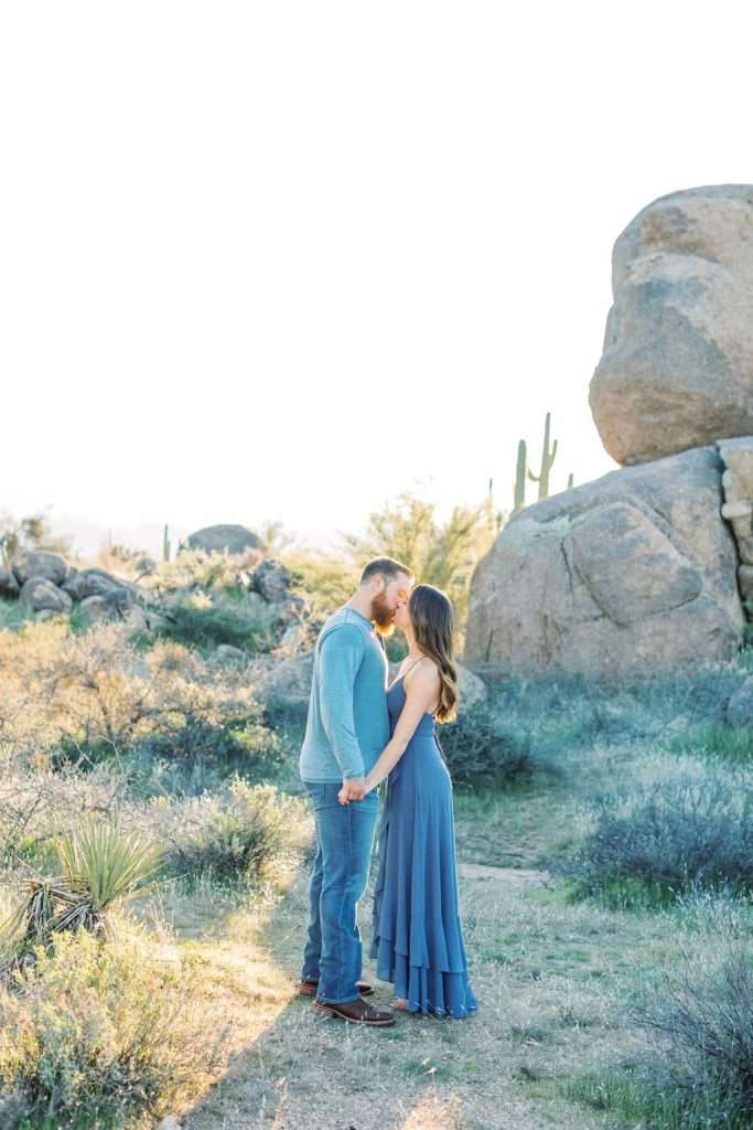 A man and woman stand holding hands and touching foreheads in a desert landscape with rocks and cacti in the background.