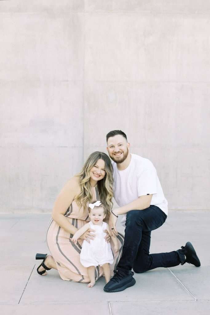 A woman, man, and baby pose together in front of a light concrete wall. The woman wears a striped dress, the man wears a white t-shirt and black pants, and the baby wears a white dress and bow.