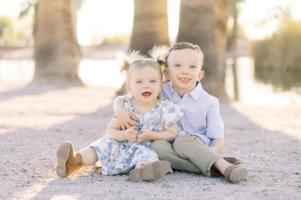 Two young children, a girl and a boy, sit on a sandy ground outdoors. The boy has his arm around the girl. They both smile at the camera with trees and sunlight in the background.