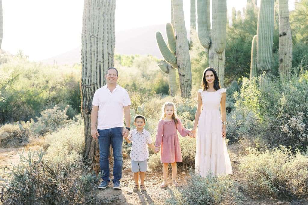 Four people, two adults and two children, stand holding hands in a desert landscape with tall cacti and shrubs in the background.