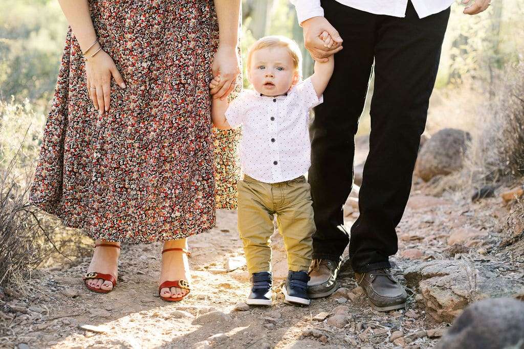 Toddler stands between two adults holding their hands while walking on a rocky outdoor path. The adults’ faces are not visible.
