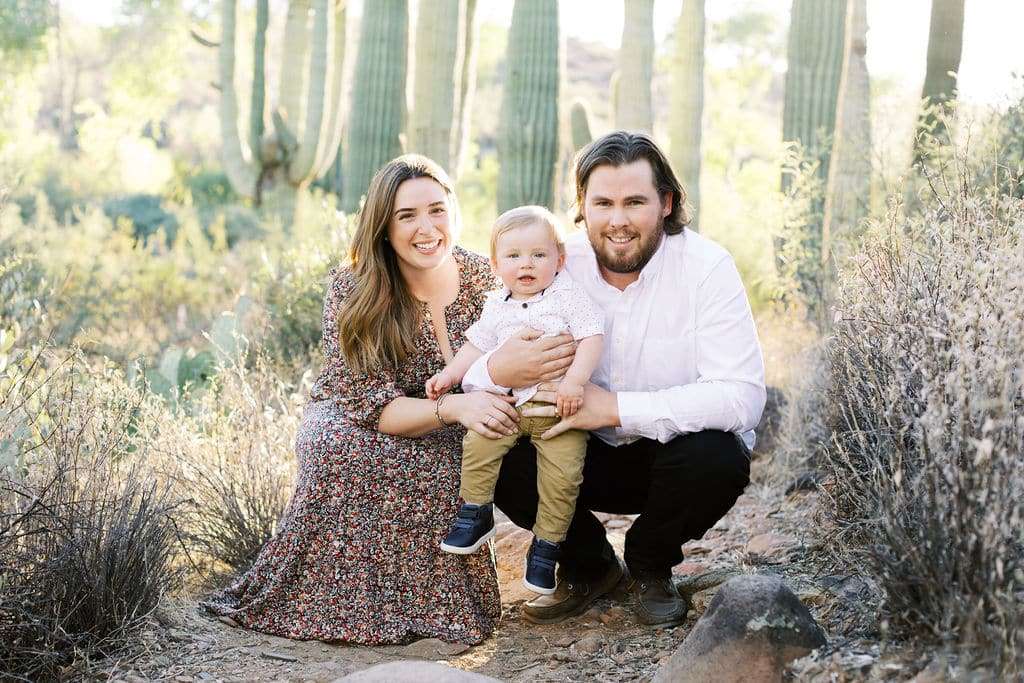 A woman, man, and toddler pose together outdoors in a desert landscape with tall cacti and dry shrubs in the background.