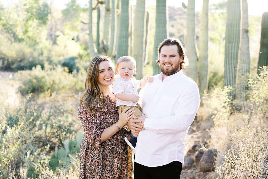 A woman and man stand outdoors holding a toddler, surrounded by tall cacti and desert plants on a sunny day.