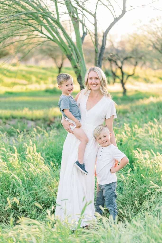 A woman in a white dress stands in a green field holding one young boy while another boy stands beside her. All three are smiling at the camera.