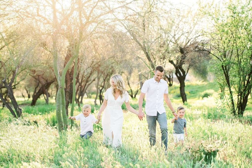A family of four walks through a sunlit grassy field, holding hands. The parents wear white, and the two young children smile and play. Trees and bright sunlight fill the background.