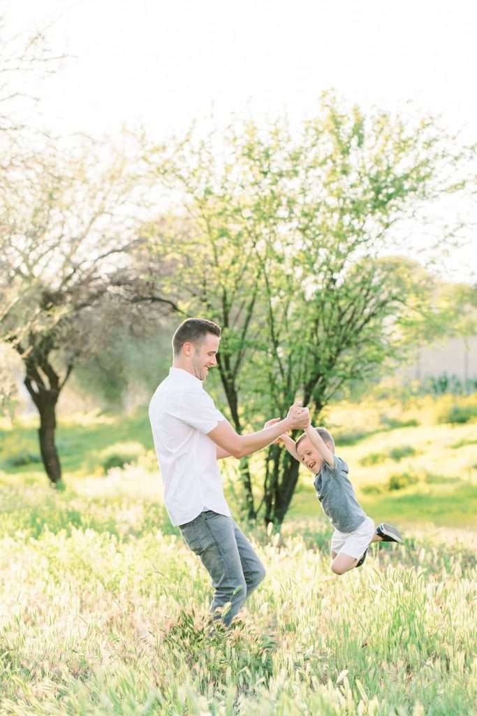 An adult spins a smiling child by the arms in a grassy field on a sunny day, with trees and greenery in the background.