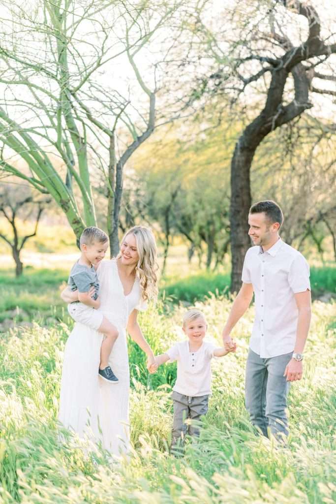 A family of four walks together in a grassy field, with the parents holding hands with their two young children on a sunny day.