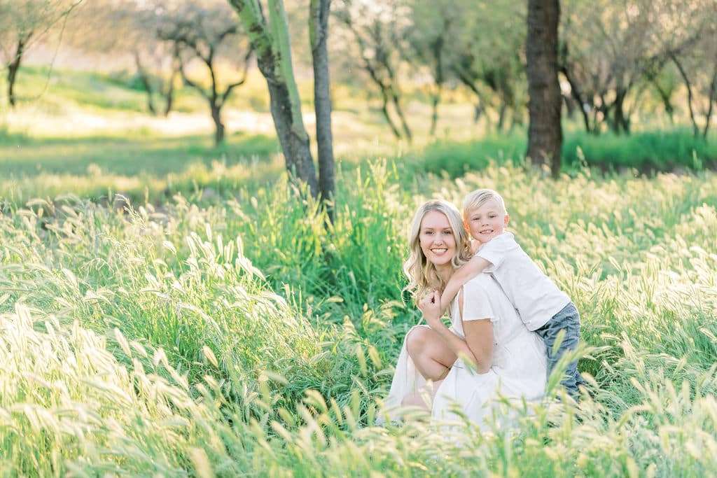 Woman in a white dress kneels in tall grass while a young boy hugs her from behind; both are smiling in a sunlit, wooded field.