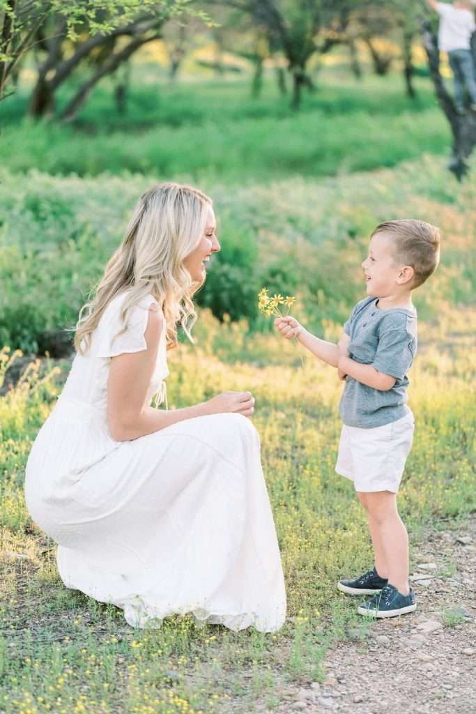 A woman in a white dress kneels on the grass while a young boy in a gray shirt and white shorts offers her a yellow flower.
