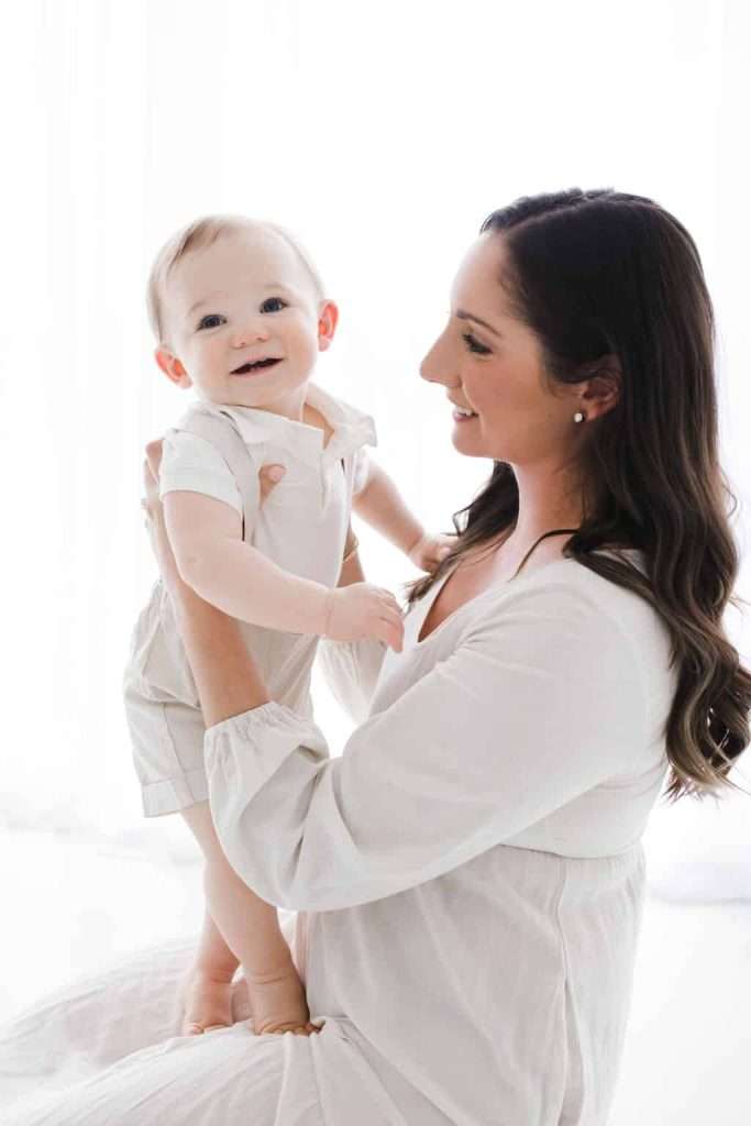 Woman holding a smiling baby dressed in white, both facing each other in a bright, softly lit setting.