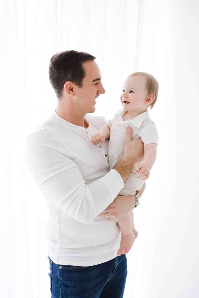 A man in a white shirt holds a smiling baby dressed in white. Both are facing each other and appear happy, standing against a bright white background.