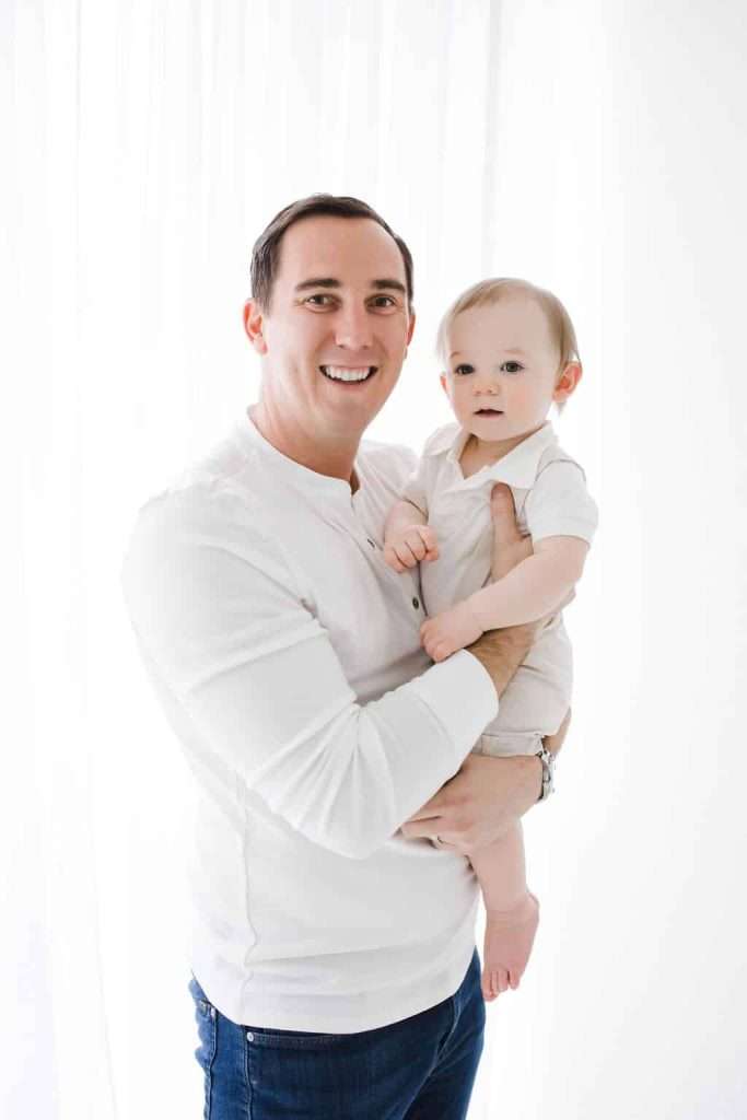 Man holding a baby, both wearing white clothes, standing in front of a bright white background.