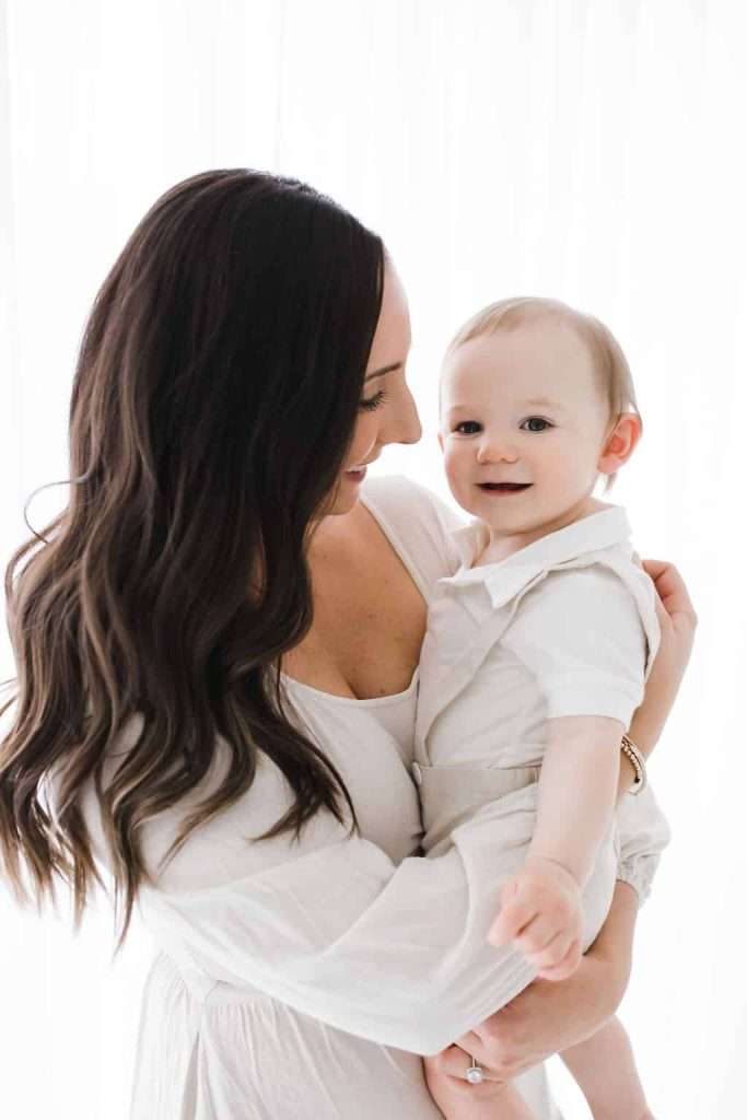 Woman with long brown hair holds a smiling baby in her arms. Both are dressed in white against a bright, softly lit background.