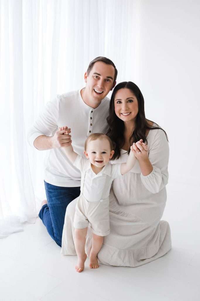 A man and woman kneel on the floor, smiling and holding hands with a toddler standing in front of them, all dressed in white clothing against a bright, white background.