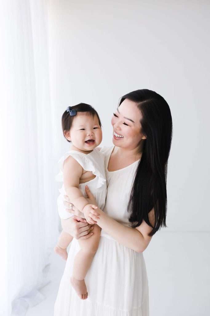 Woman in a white dress holding a smiling baby with a hair bow, both facing the camera, in a bright, minimalist room.