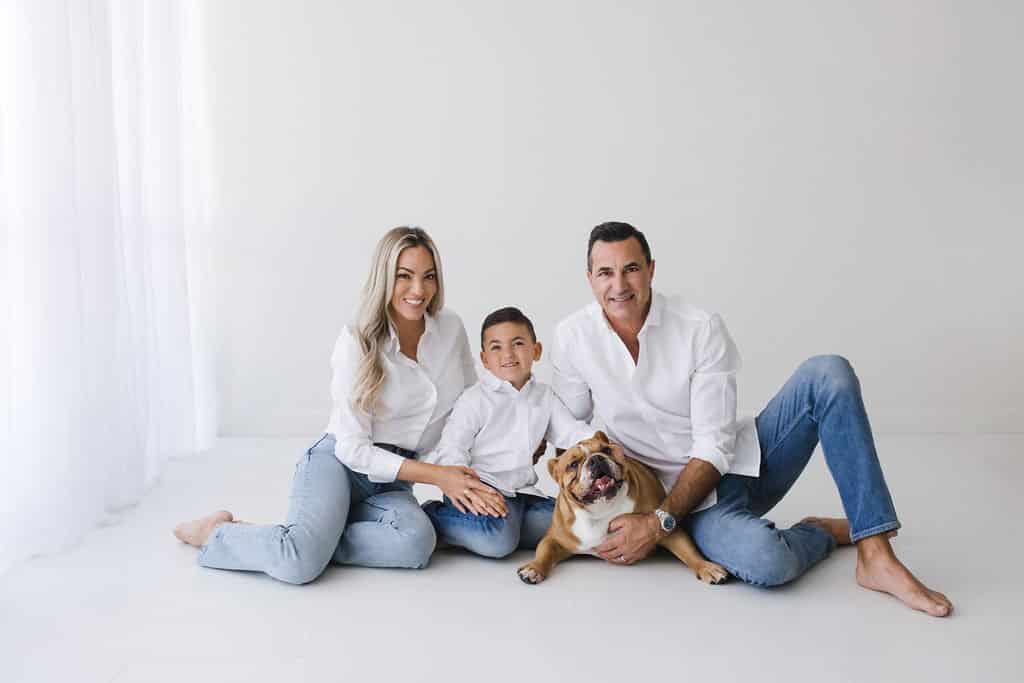 A woman, man, boy, and bulldog sit on the floor against a white background, all wearing white shirts and blue jeans, smiling at the camera.