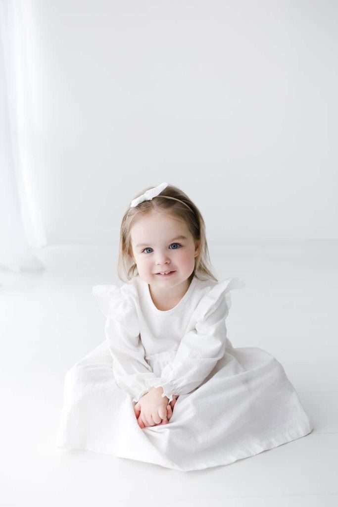 Young child with light brown hair and a white bow sits on the floor wearing a long-sleeved white dress, looking at the camera in a bright, minimalist setting.