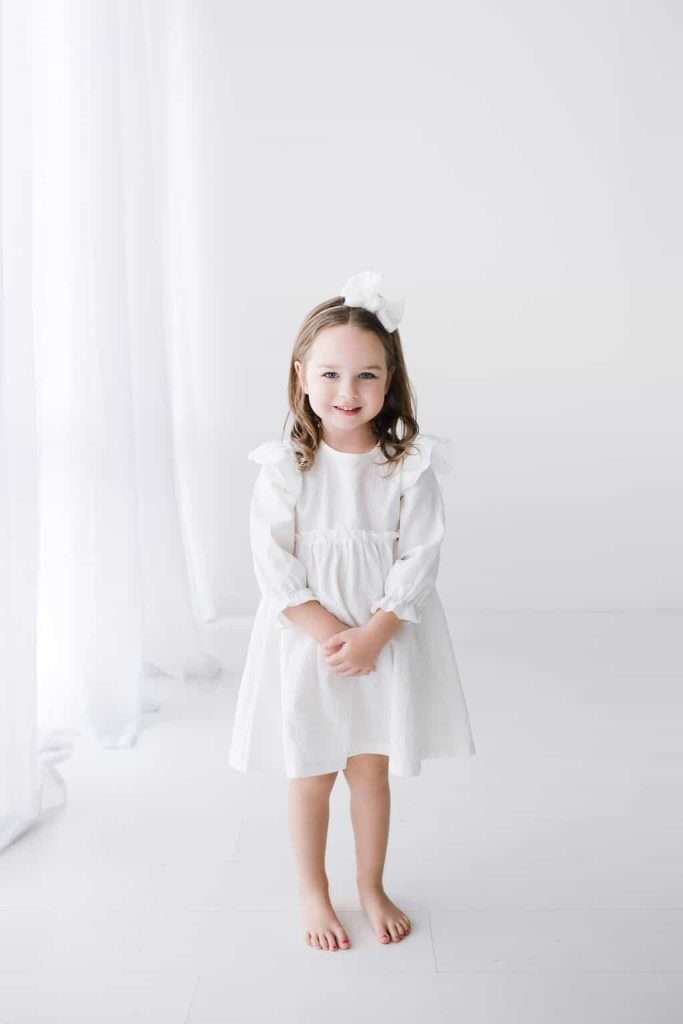 Young girl with light brown hair stands barefoot in a white dress with a large white bow in her hair, posing against a white background with sheer curtains.