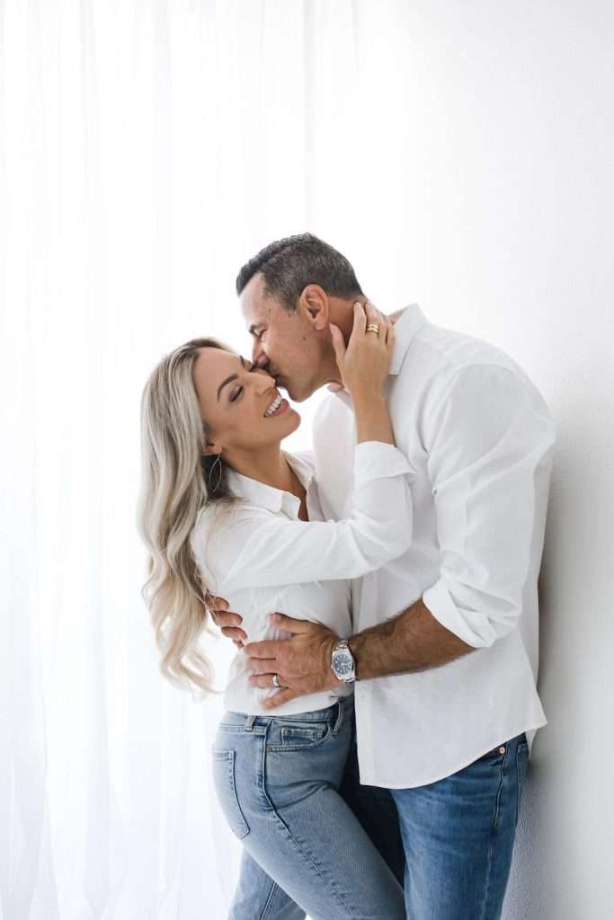 A couple dressed in white shirts and jeans stands close together, smiling and embracing in front of a white background.