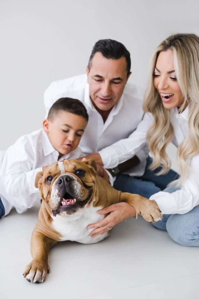 Smiling family with a young boy and a woman petting a happy bulldog on the floor, all wearing white tops and posing together indoors.
