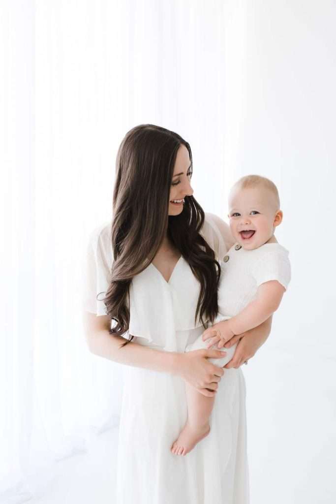 Woman with long brown hair holds a smiling baby dressed in white. Both are looking at each other and standing in a bright, white room.
