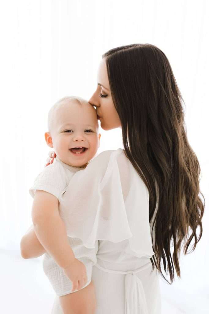 Woman with long brown hair holds and kisses a smiling baby on the forehead. Both are dressed in light-colored clothing, standing against a bright, white background.