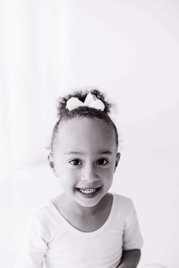 Young child with curly hair and a white bow smiles at the camera, wearing a light-colored shirt, against a plain background.