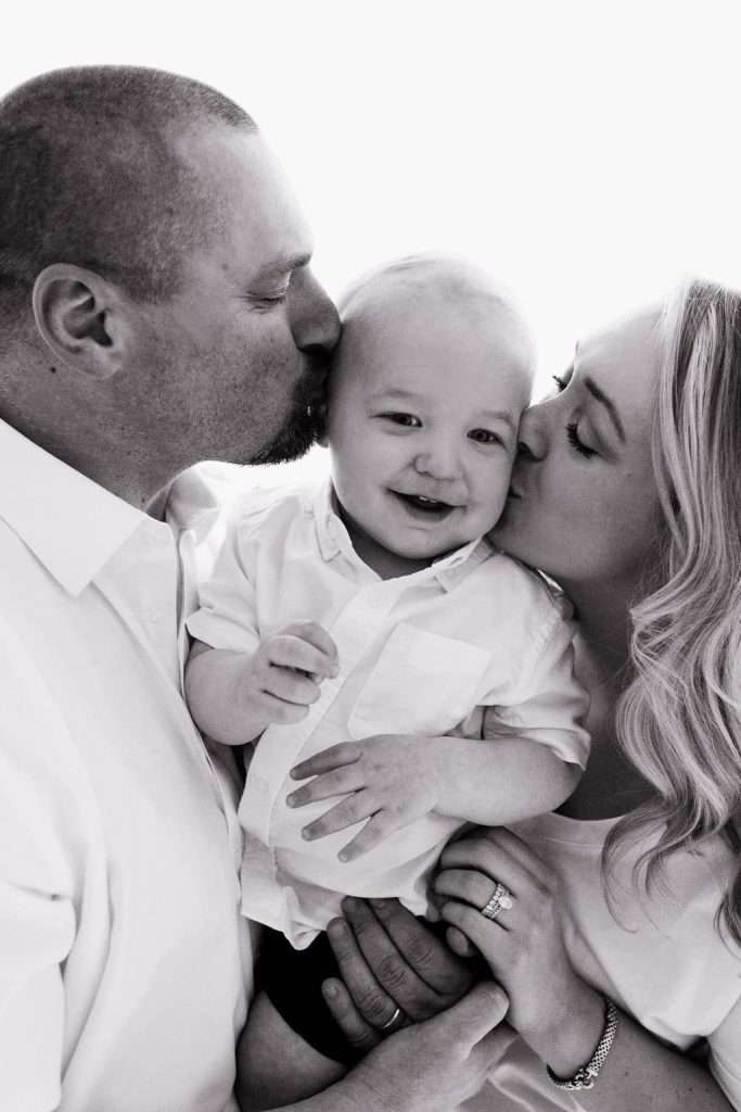 Black and white photo of a man and woman kissing a smiling baby on the cheeks; all three are wearing light-colored shirts.