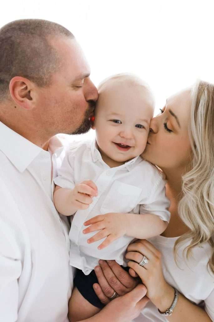 A man and woman kiss a smiling baby on the cheeks. All three wear white shirts and are closely posed for the photo.