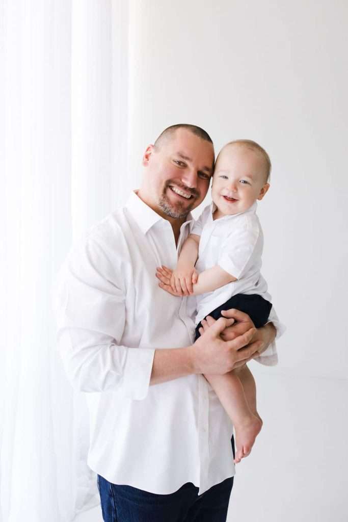 Man holding a smiling baby, both wearing white shirts, standing in a bright, white room with sheer curtains.