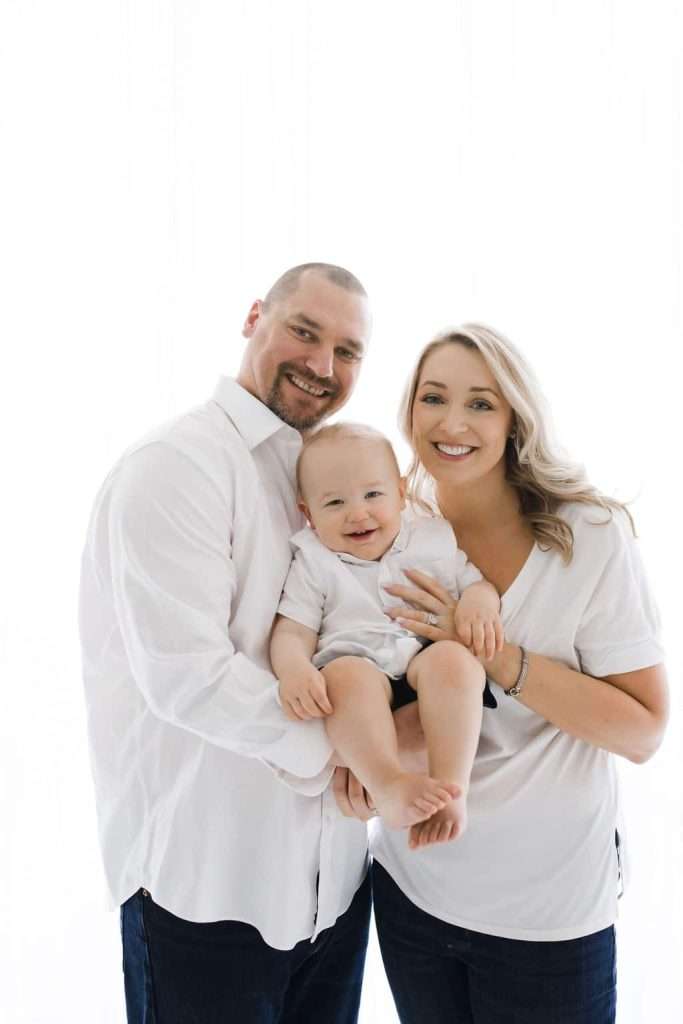 A man and woman stand close together, smiling and holding a baby. All three wear white tops and dark pants against a bright white background.