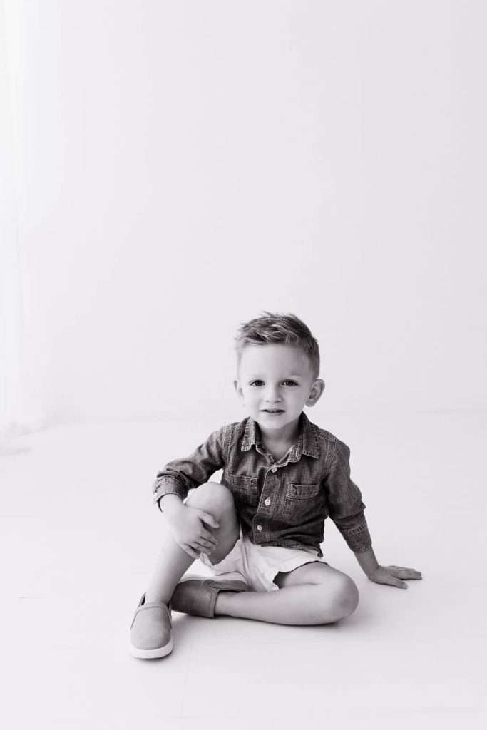 Young boy sitting on the floor with one knee up, wearing a denim shirt and shorts, looking at the camera. Black and white photo.
