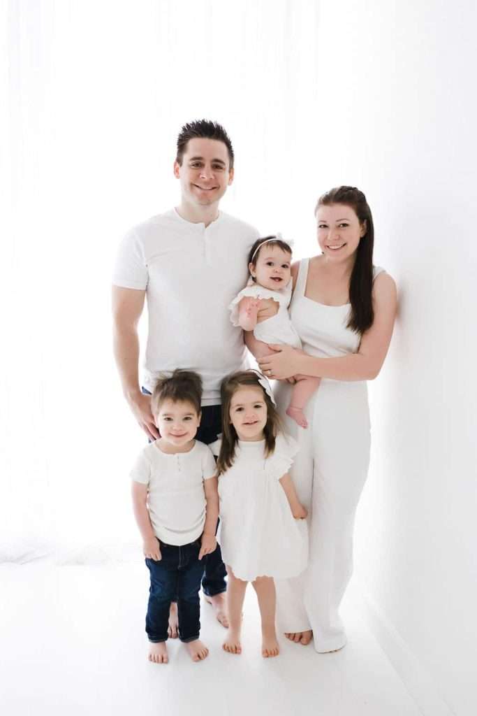 A family of five, all dressed in white, poses against a white background. The parents stand behind, with the mother holding a baby, and two young children stand in front.