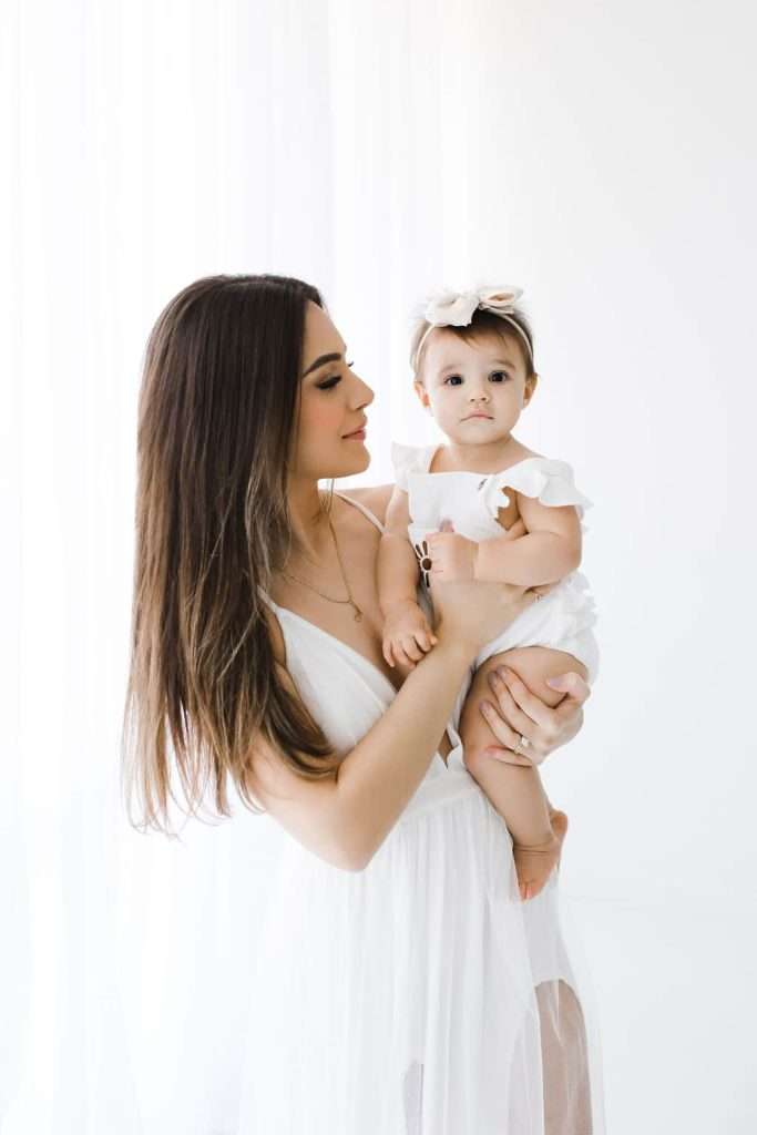 A woman in a white dress holds a baby girl dressed in white with a headband, standing against a white background.
