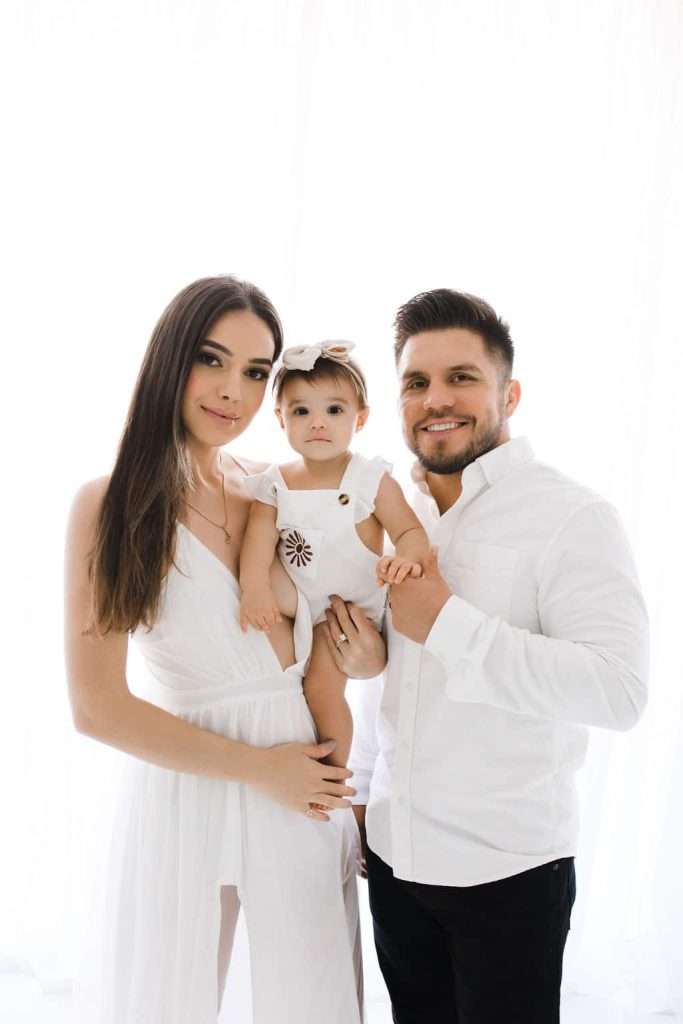 A woman and man stand together holding a baby girl. All three wear white outfits and pose against a bright, white background.