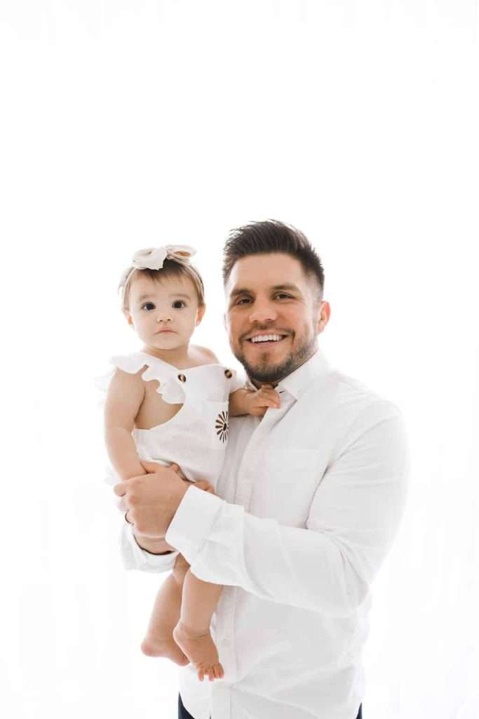 Man in a white shirt holds a baby wearing a white outfit and bow, both facing the camera against a plain white background.