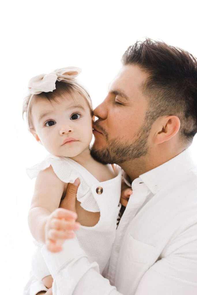 A man in a white shirt holds a baby girl wearing a white outfit and bow, kissing her on the cheek.