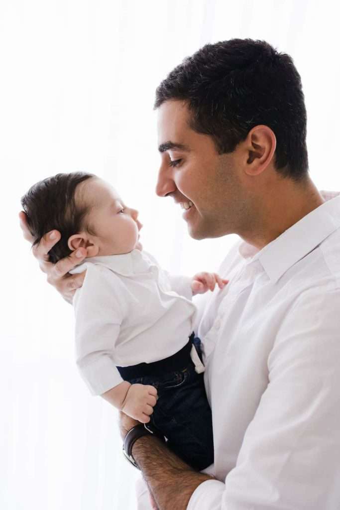 An adult holds a baby close, both dressed in white shirts. The adult smiles and gently touches noses with the baby against a light background.