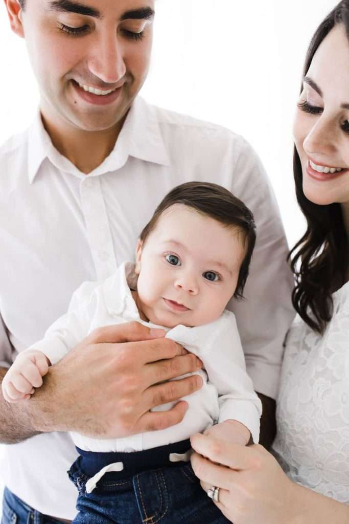 A man and woman, both in white shirts, hold a baby with dark hair and blue eyes, who is looking at the camera.