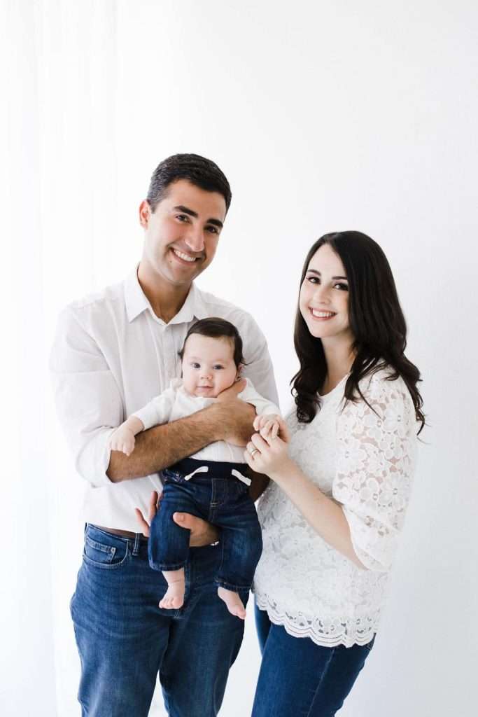 A man and woman, both in white tops and blue jeans, stand closely together holding a baby dressed in a light shirt and jeans, all smiling against a plain white background.