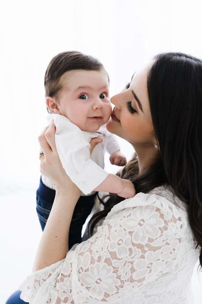 Woman holding a baby in her arms, gently touching her nose to the baby’s cheek. Both are wearing white clothing, and the background is bright and minimal.