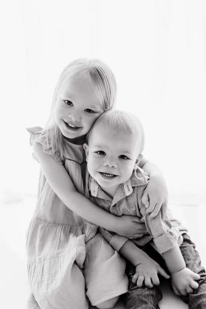 Two young children, a girl and a boy, sit together and smile at the camera. The girl has her arm around the boy in a friendly, affectionate pose. The photo is in black and white.