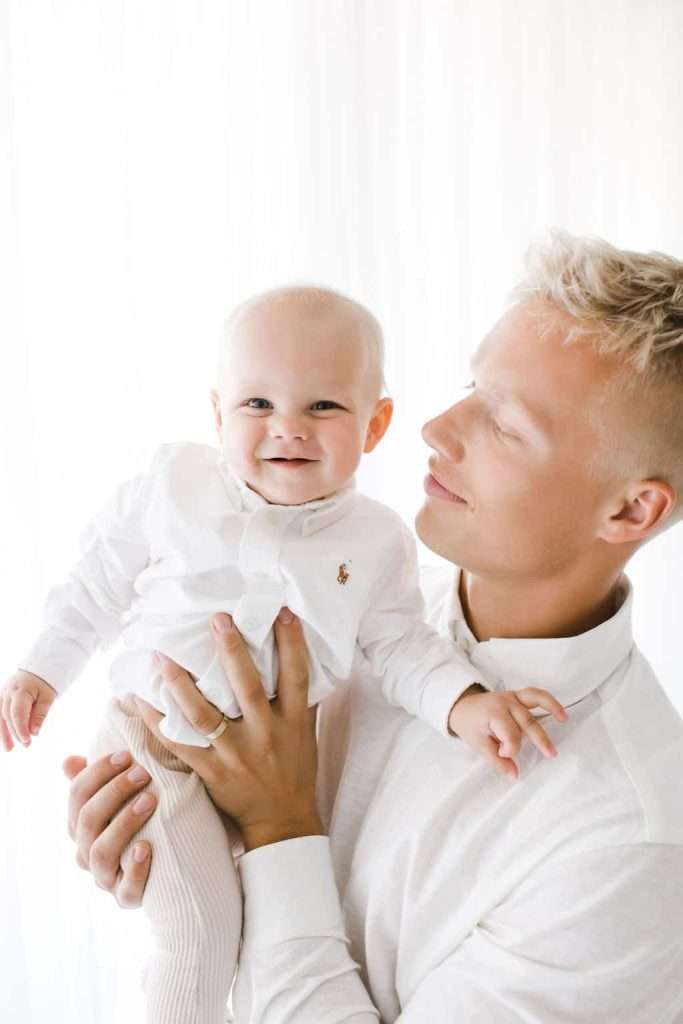 A man in a white shirt holds a smiling baby dressed in a white outfit against a bright, white background.