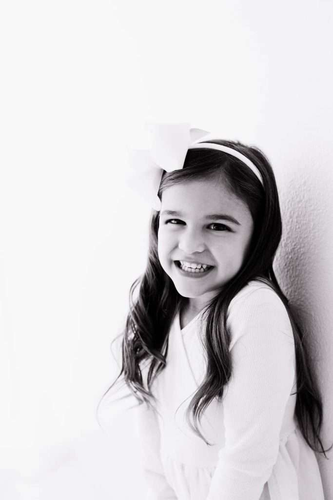 Smiling young girl with long hair and a large bow on her head, wearing a light-colored dress, standing against a plain wall. Black and white photo.